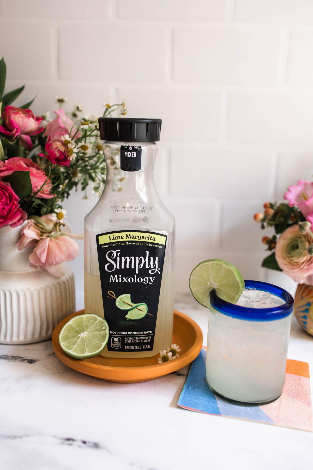 A big bottle of Simply Mixology with a sliced lime beside it on an orange plate next to a blue-rimmed margarita glass garnished with a lime wedge.