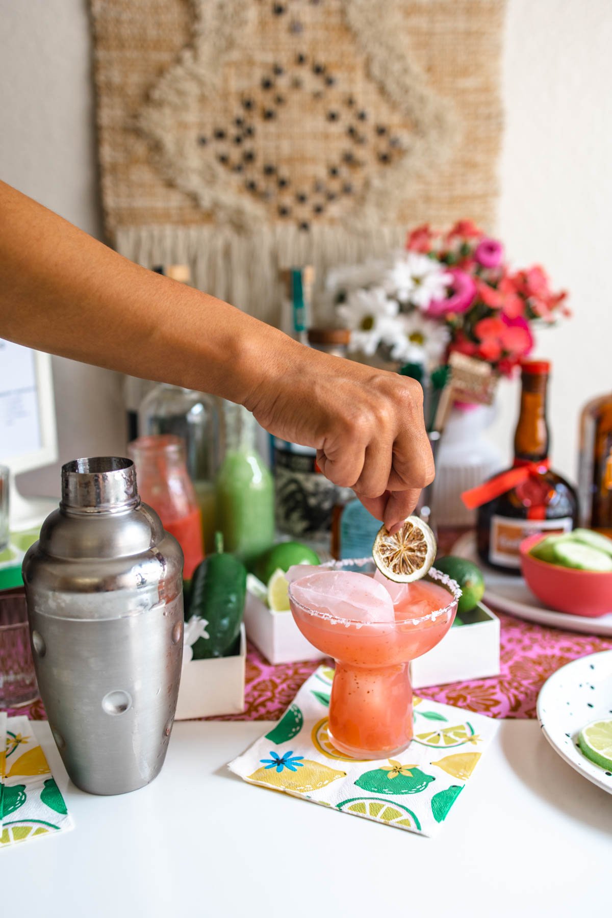 A person placing a dried lime wheel on top of a strawberry margarita that's beside a cocktail shaker and in front of various margarita ingredients and supplies.