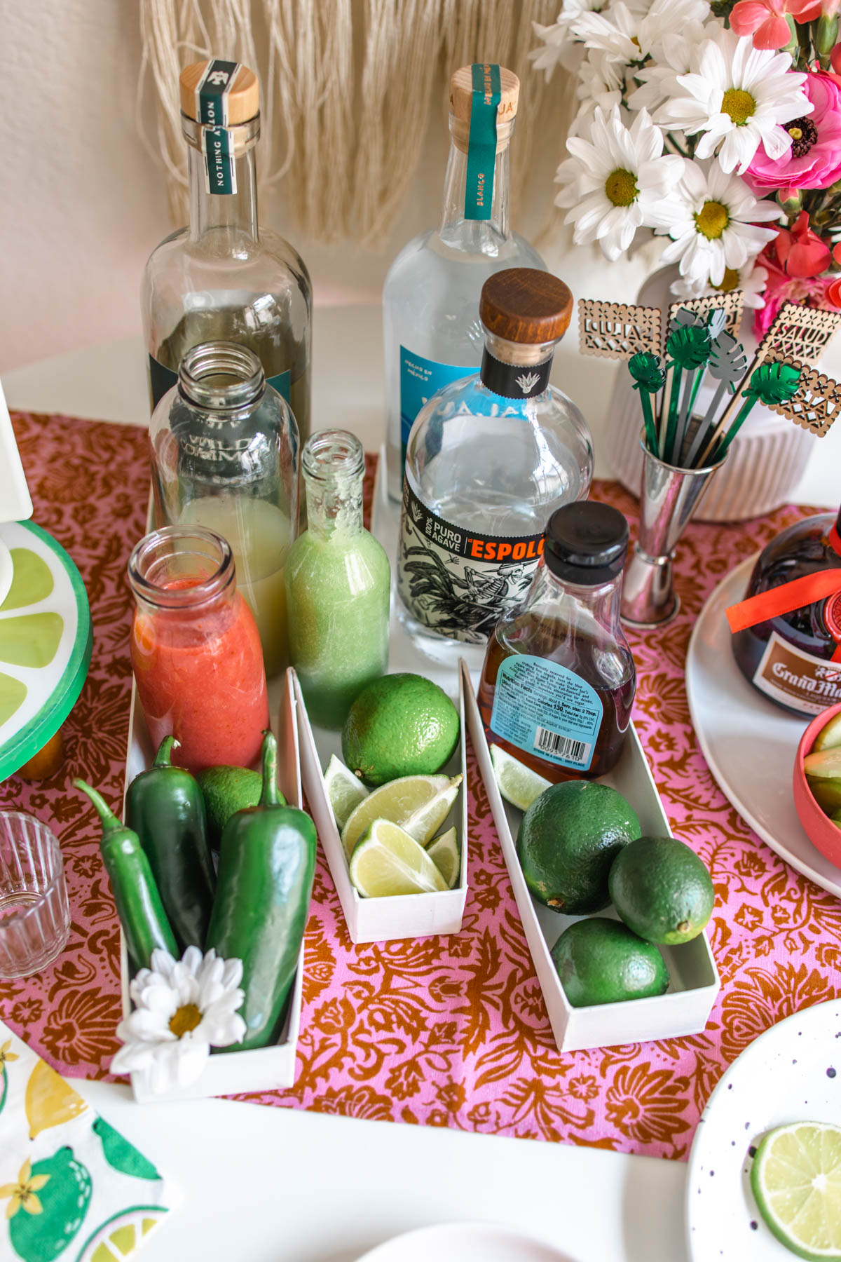A photo of tequila bottles, agave, simple syrup, and cucumber and strawberry purees along with whole lime and peppers as well as sliced limes inside rectangular boxes.