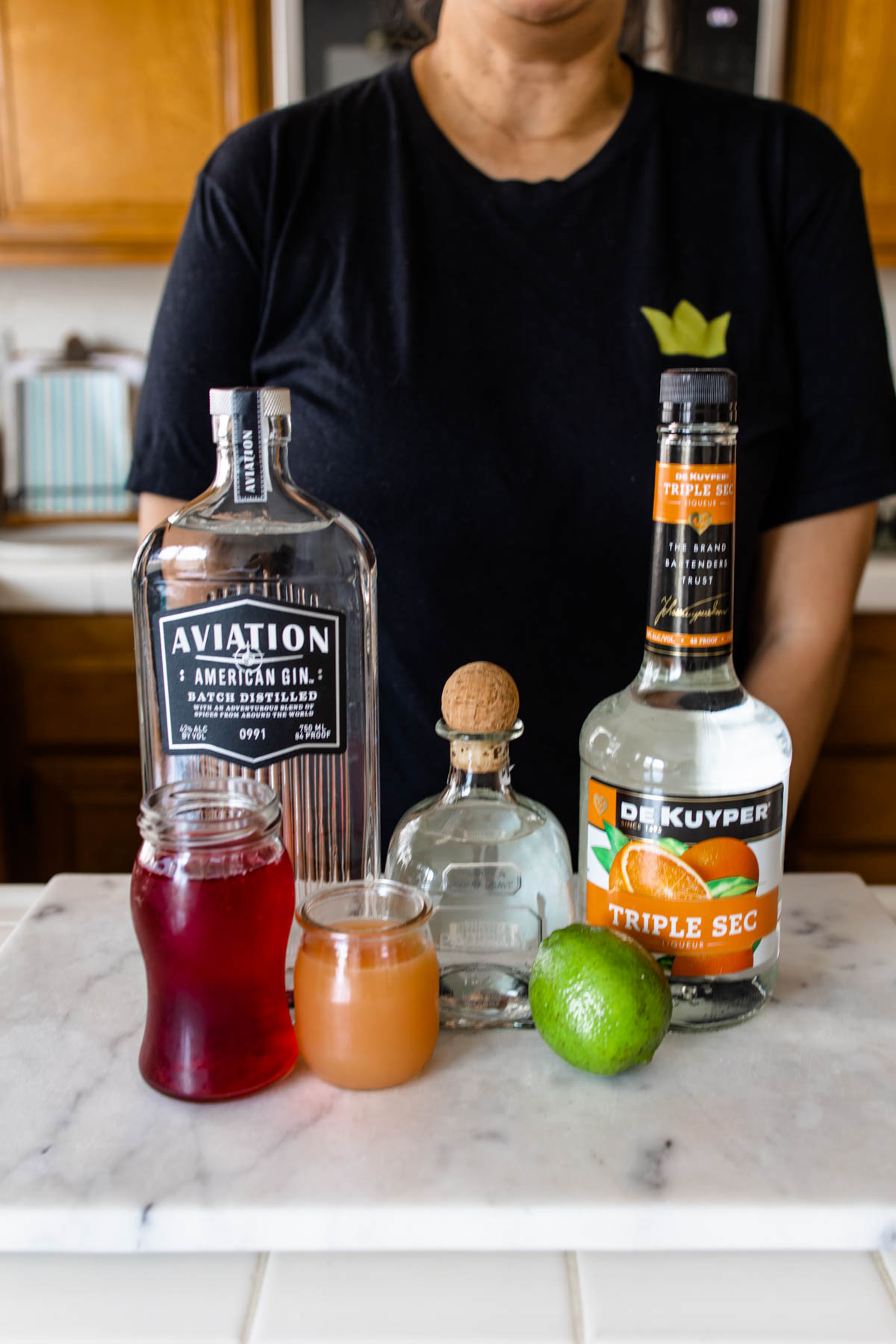 Bottles of Aviation Gin, Patron Silver tequila, and Triple Sec orange liqueur behind containers of cranberry and grapefruit juice and a fresh lime on top of a counter.