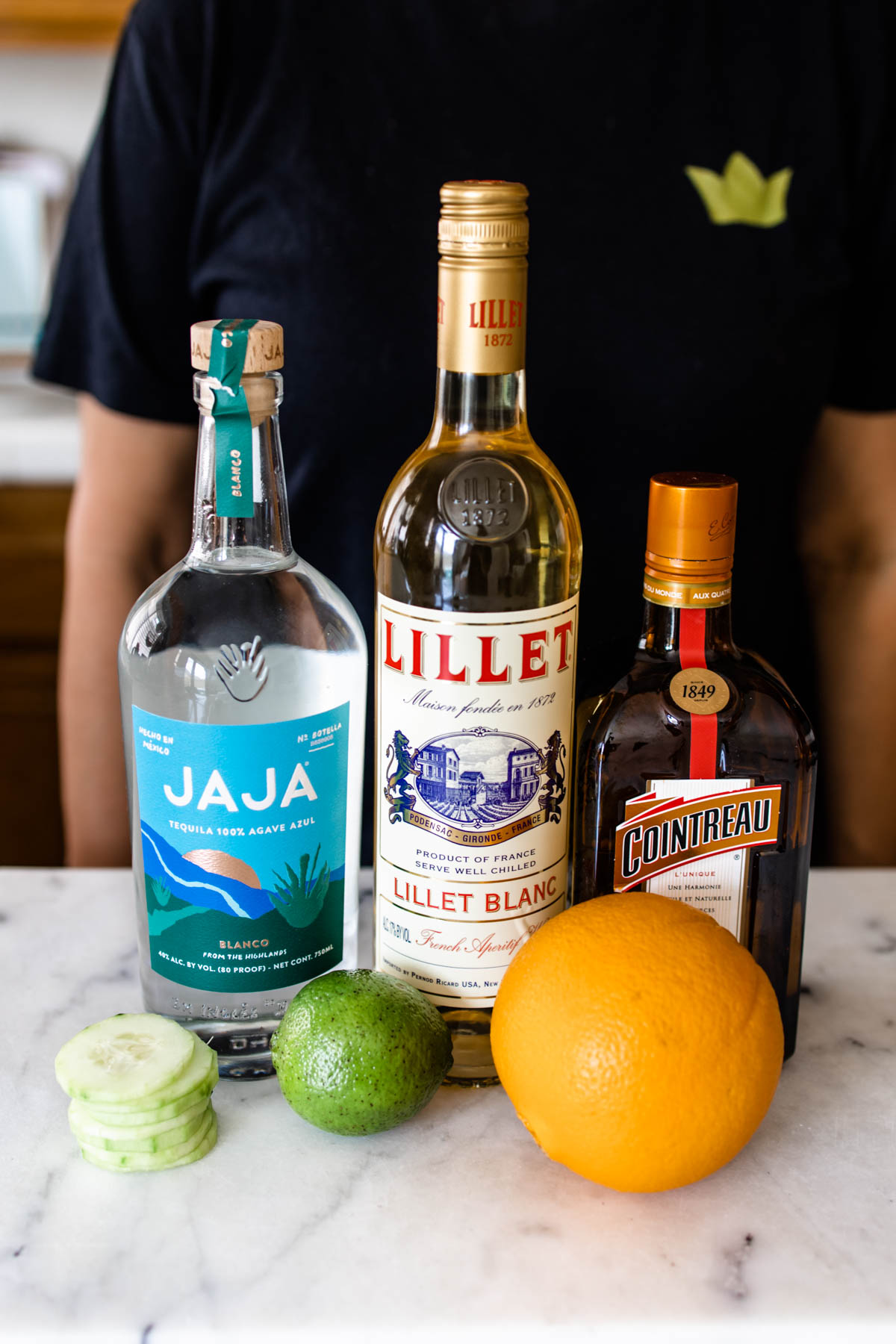 Bottles of JAJA Tequila, Lillet Blanc, Cointreau, along with lime and orange on a white counter.