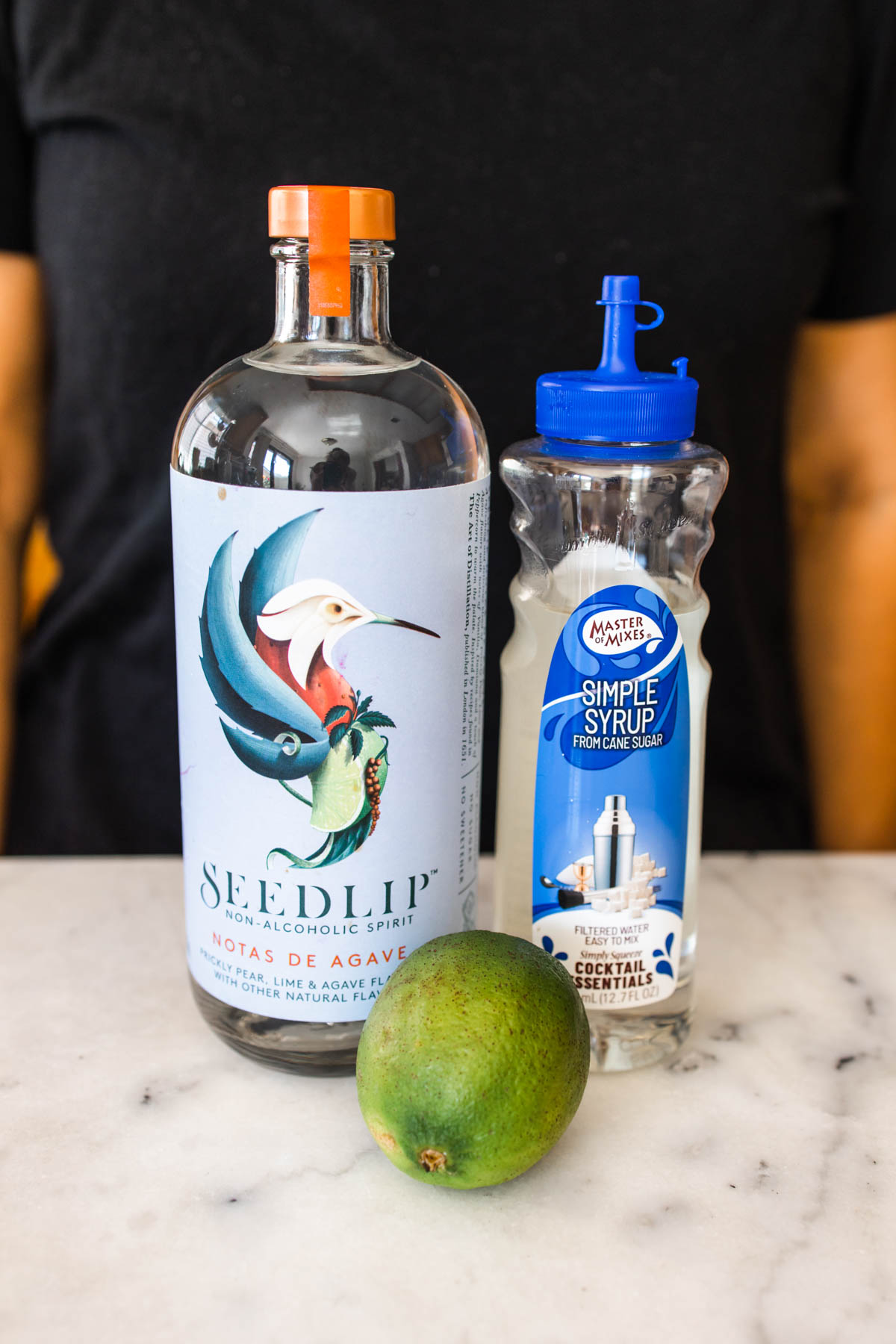 A bottle of Seedlip Notas de Agave beside a container of Master Mixes Simple Syrup with fresh lime in between them on top of a counter.