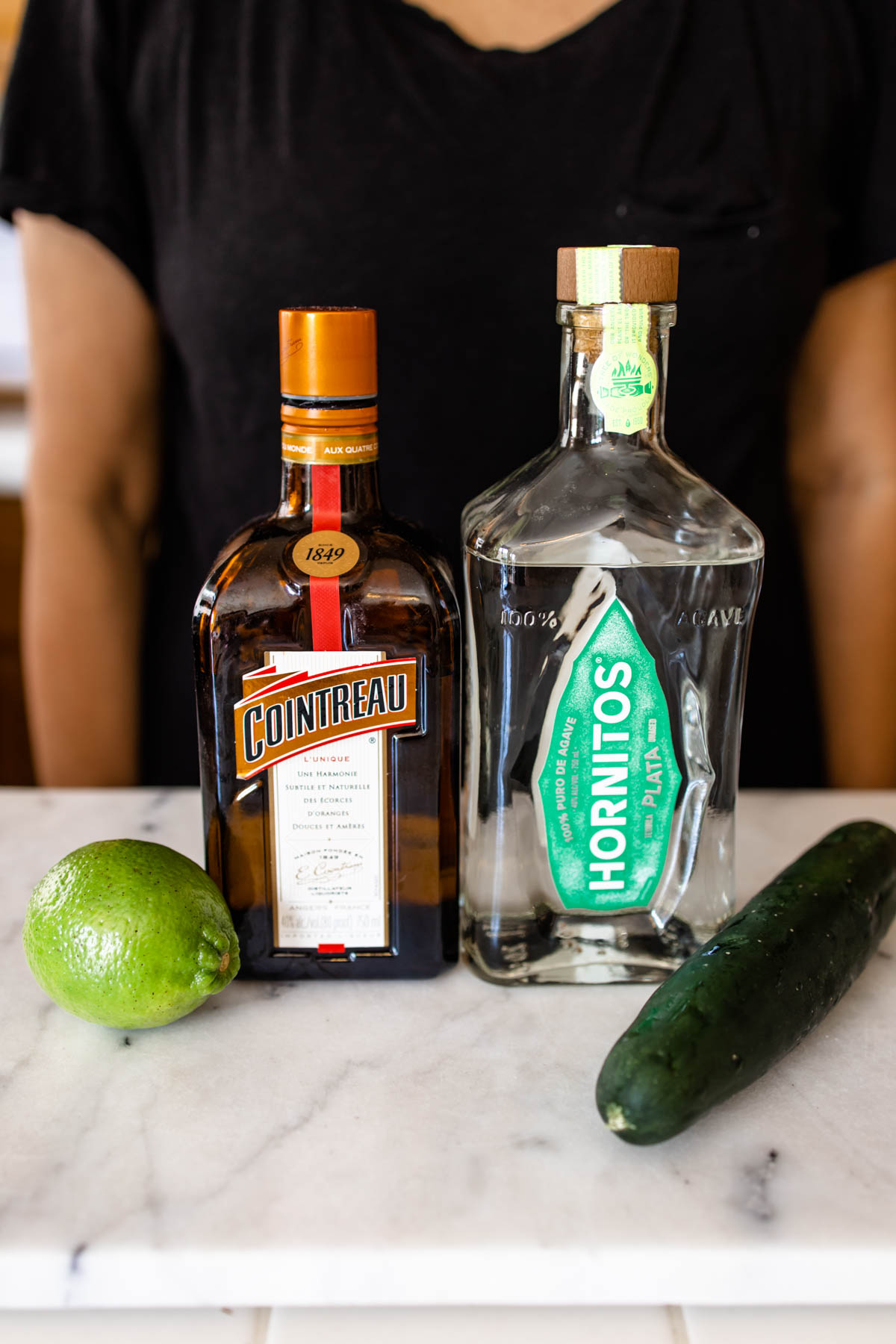 Bottles of tequila and Triple Sec in between a fresh lime and fresh cucumber on a marbled countertop.