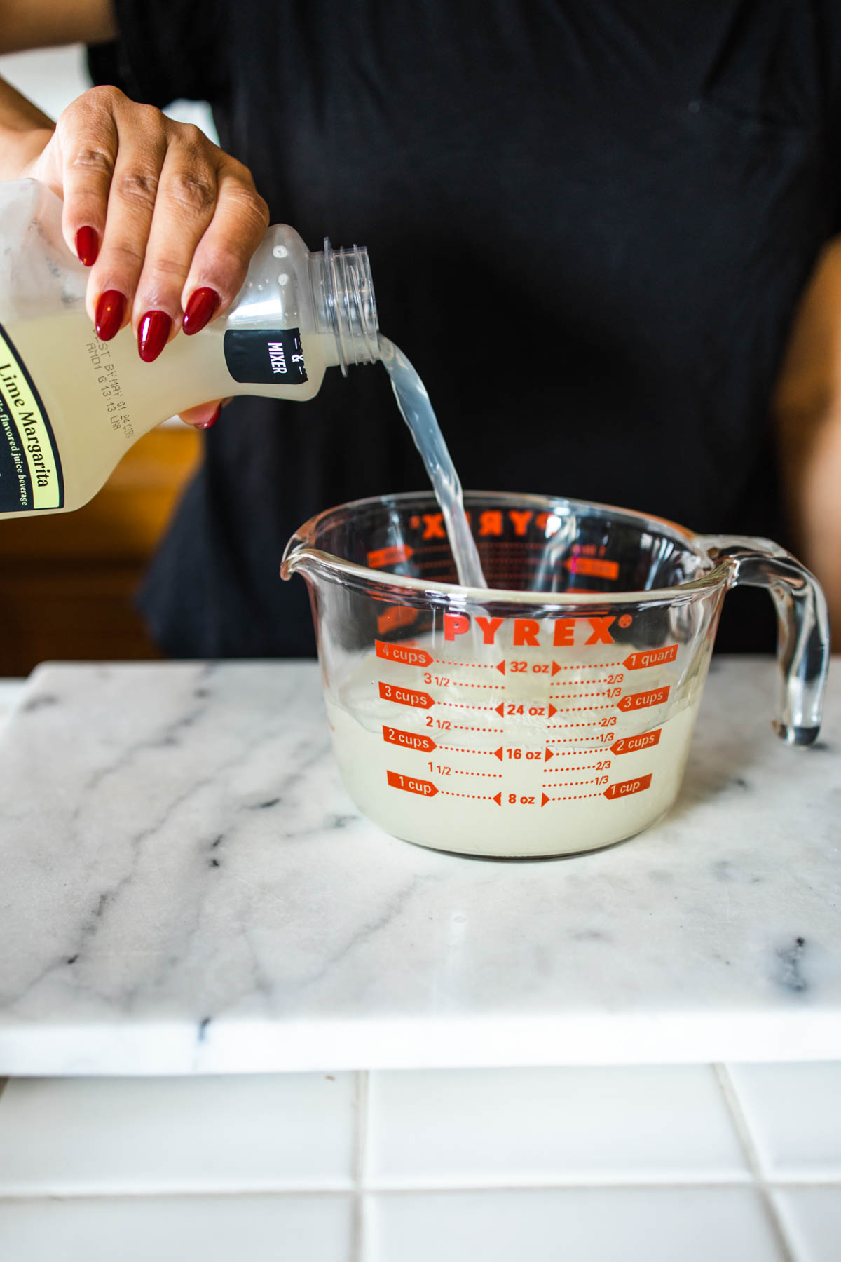 A person pouring some Simply Mixology Lime Margarita to a measuring cup on a white counter.