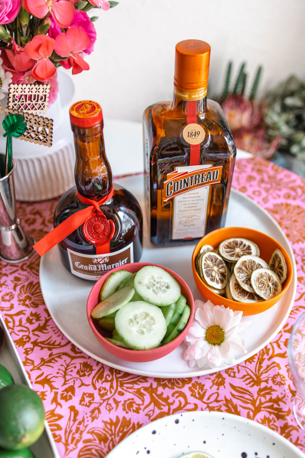 Bottles of Grand Marnier and Cointreau behind two bowls filled with cucumber slices and dried lime wheels.