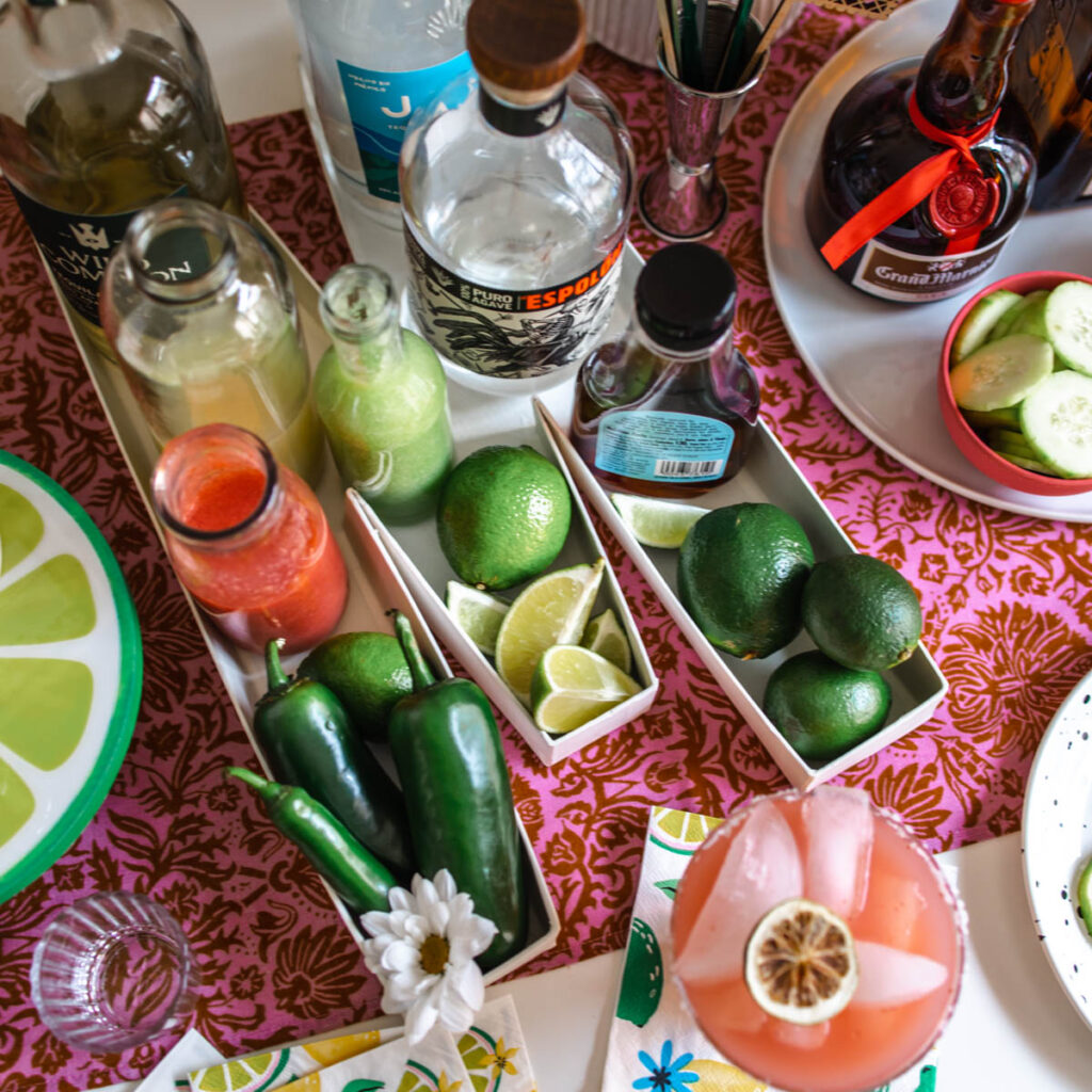 An image of tequila bottles, Grand Marnier, fresh jalapenos and limes, and a strawberry margarita with dried lime wheel at the bottom.