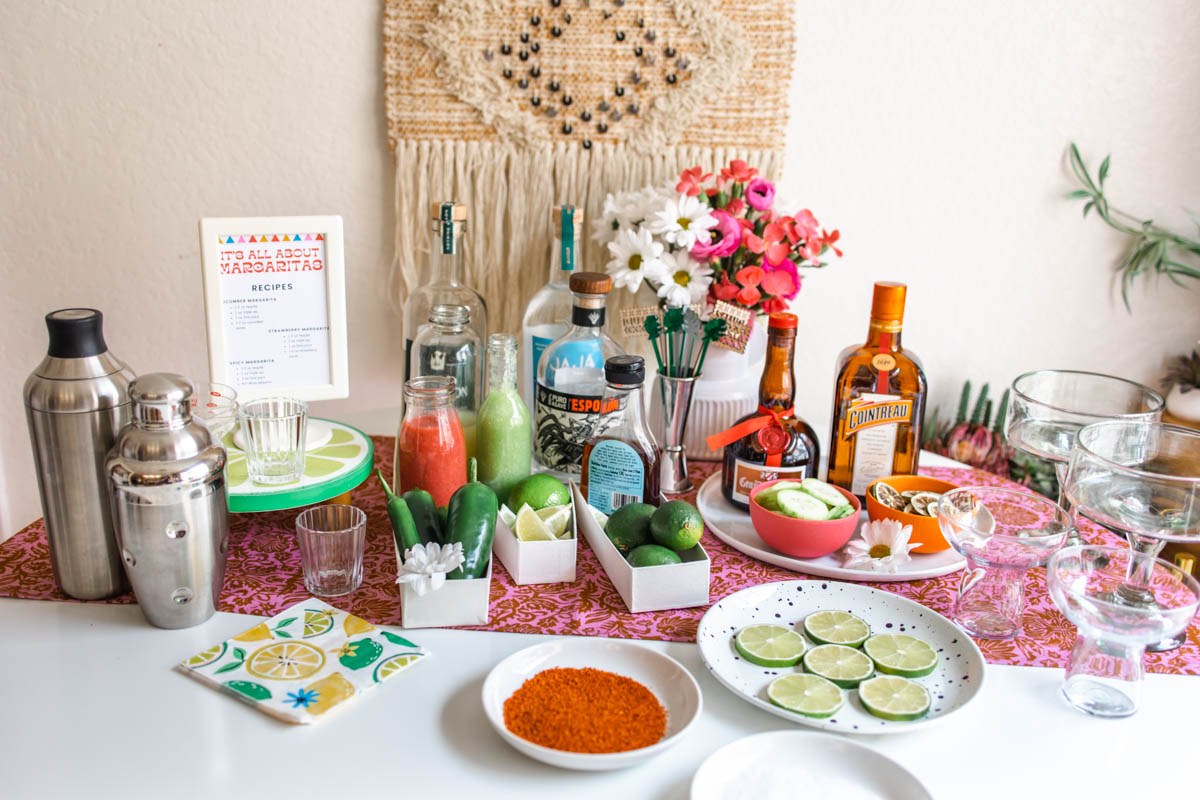A margarita bar set up with cocktail shakers, margarita recipes, tequila and orange liqueur bottles, fruit juices, peppers, glasses, and plates with Tajin and sliced limes.