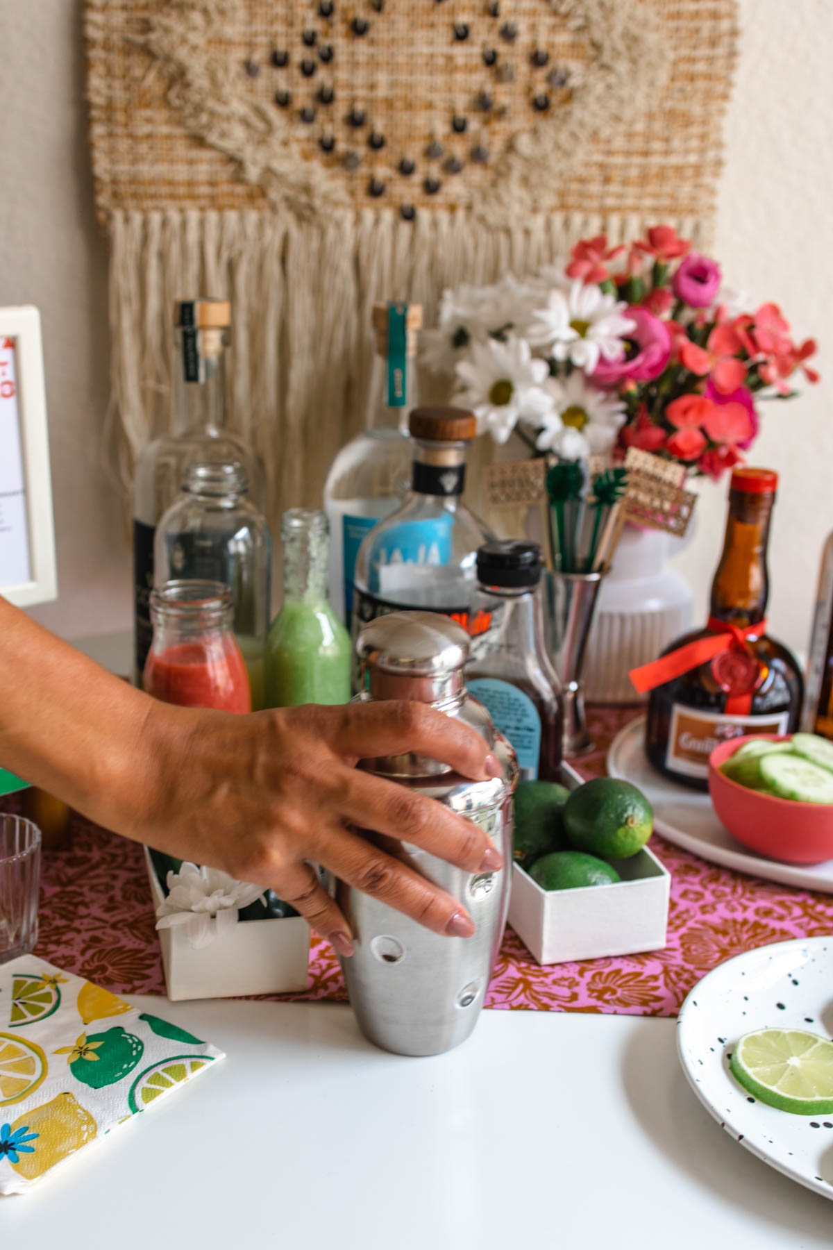 A person holding a cocktail shaker in front of margarita ingredients and supplies on a table.