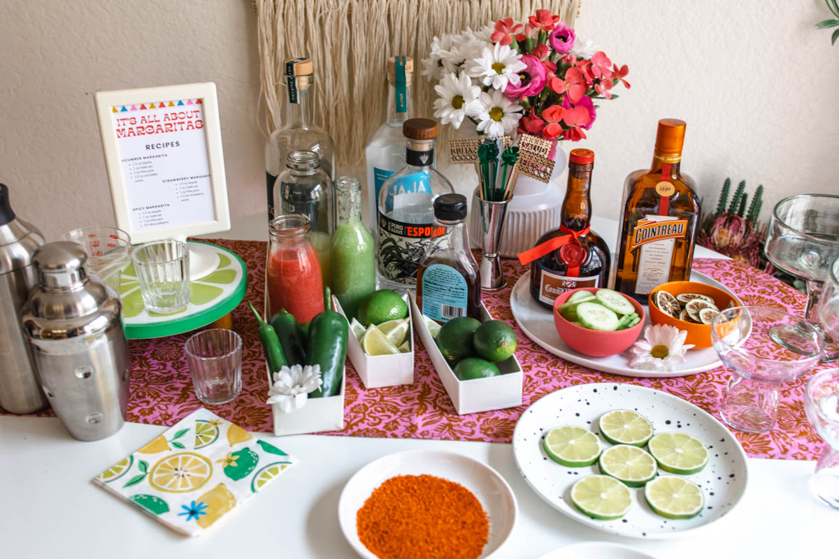 A margarita bar full of ingredients, equipment, and supplies for making margaritas including cocktail shakers, bottles of tequila, orange liqueur, and fruit juices, fresh lime and jalapenos, and cut limes and Tajin on plates.