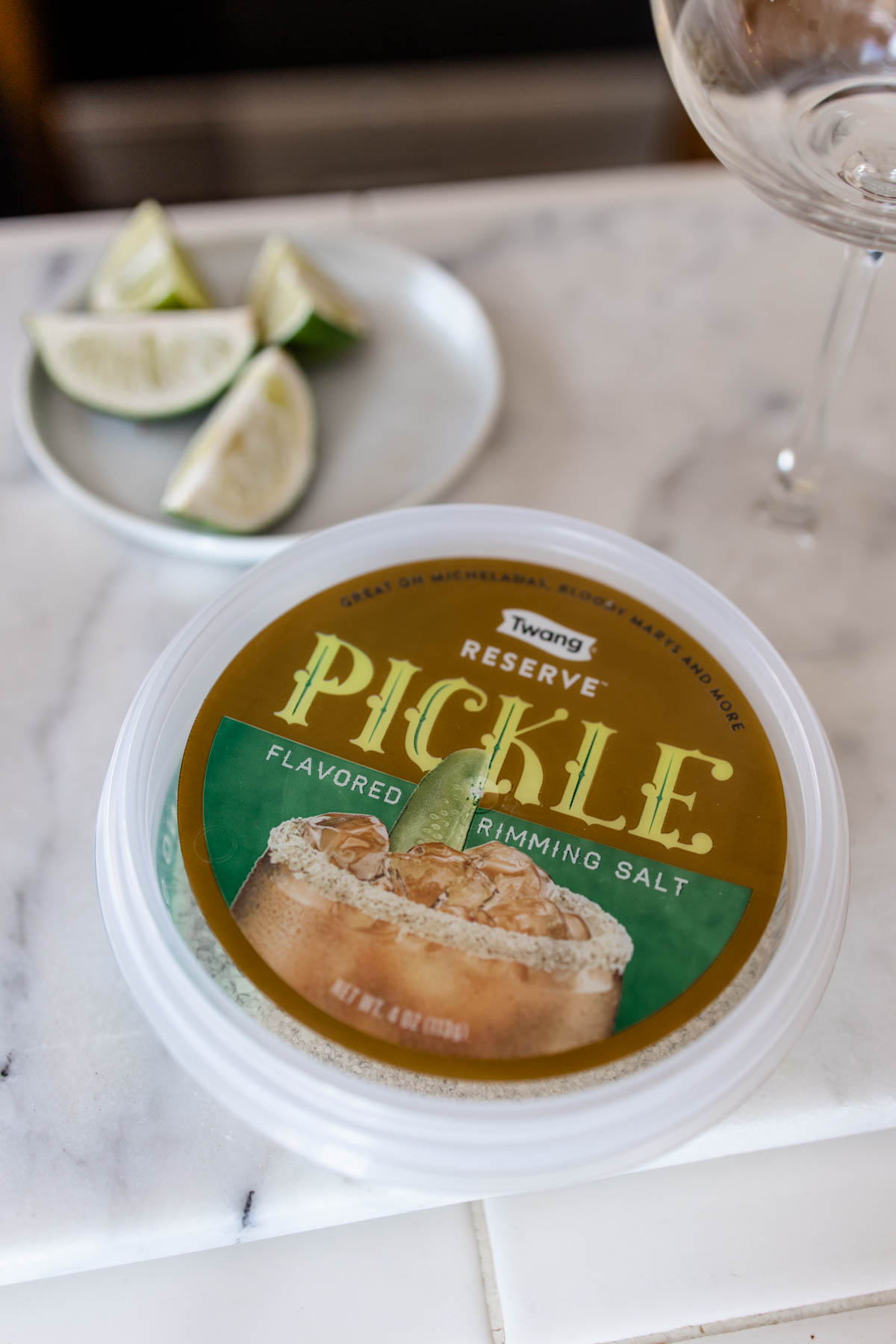 A container of Twang Reserve Pickle Flavored Rimming Salt with a plate of lime wedges behind it on a counter.