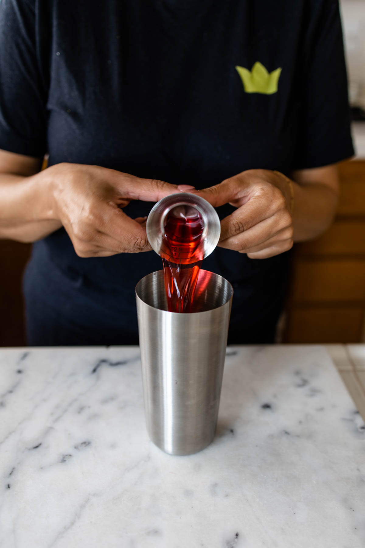 A person pouring some red cranberry juice into a cocktail shaker using a jigger.