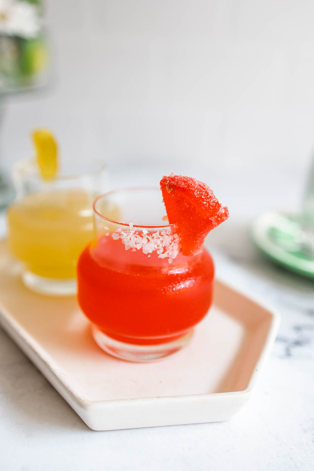 A deliciously red Deadpool Margarita in a glass with salt rim garnished with a candy watermelon slice alongside a yellow Wolverine Margarita on a plate.  