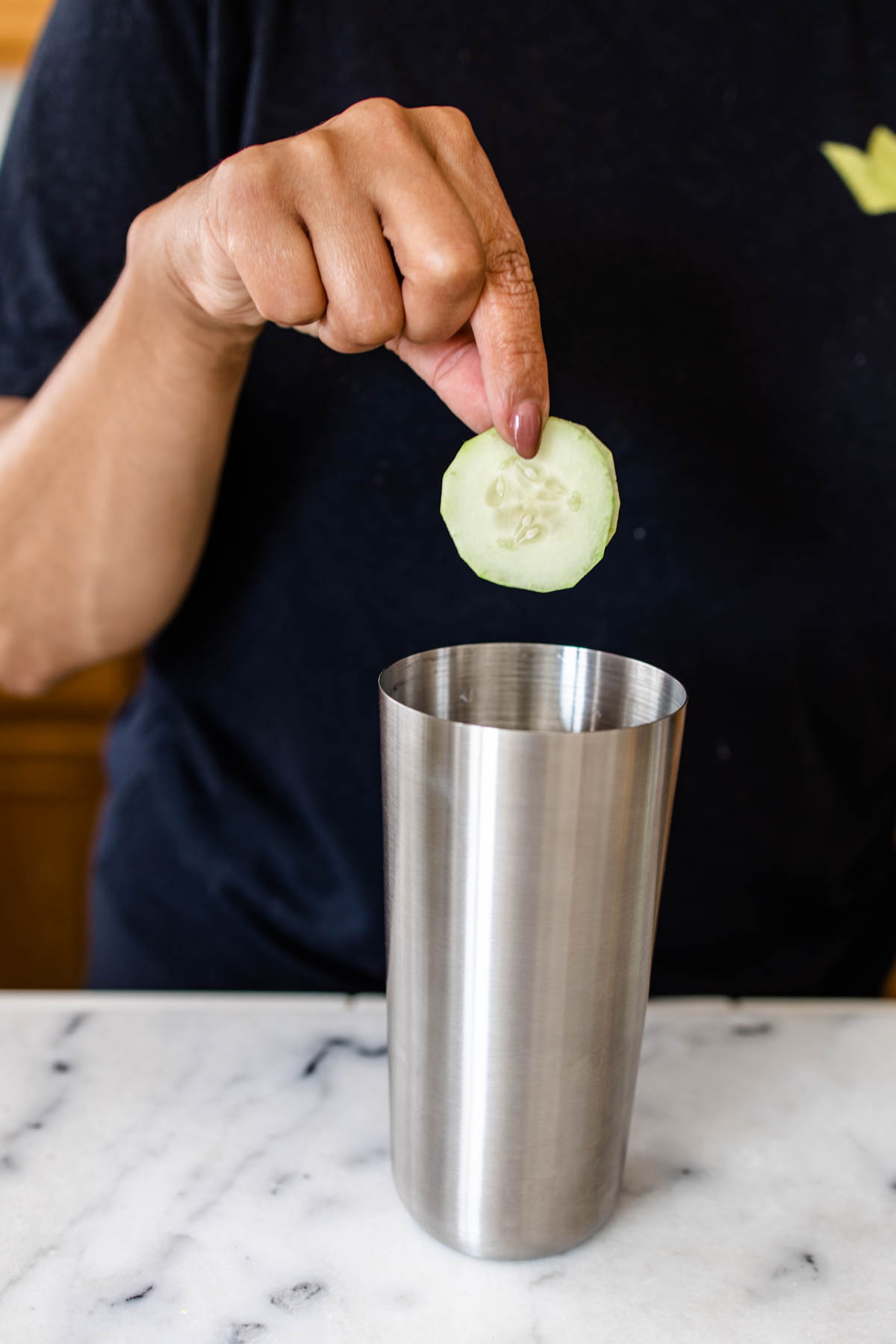 A person adding a cucumber slice into a cocktail shaker.