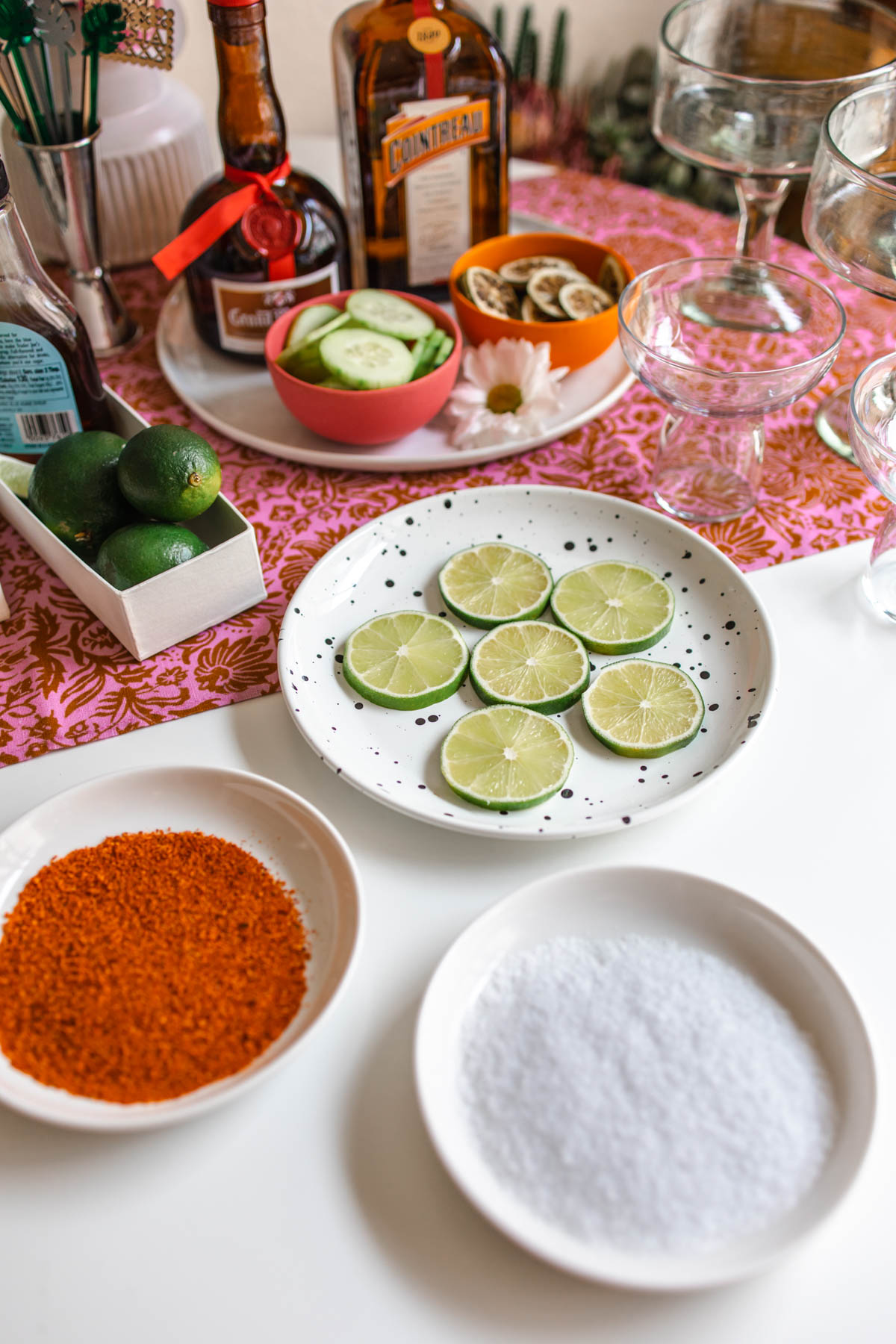 Plates of salt, Tajin, and sliced limes in front of bottles of orange liqueurs and bowls of cucumber slices and dried lime wheels.