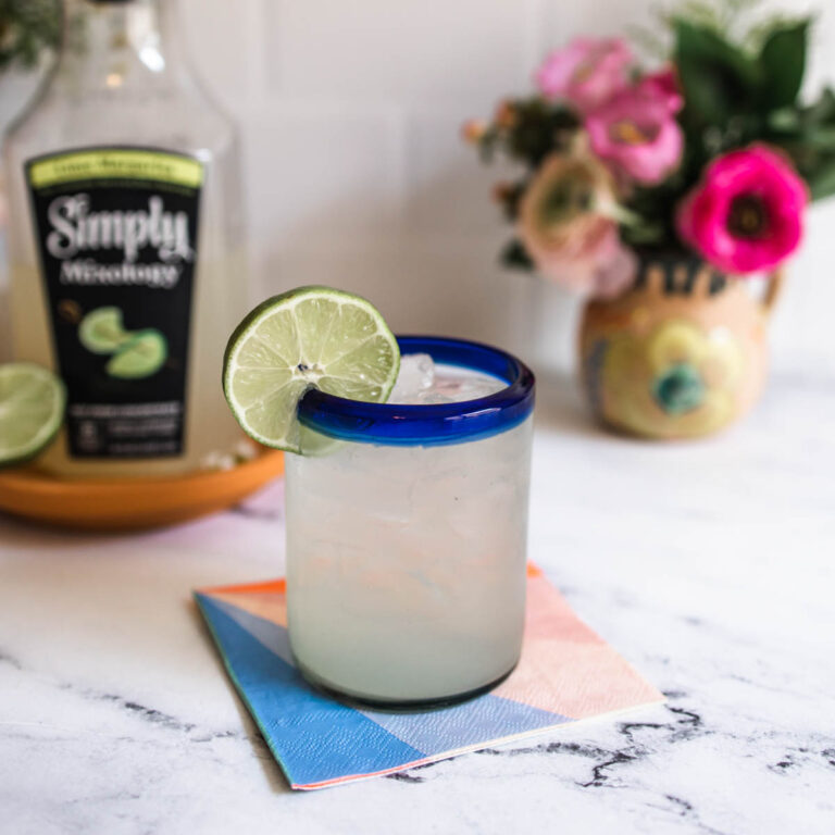 A full blue-rimmed glass of margarita garnished with a lime wedge in front of a bottle of Simply Mixology Lime Margarita. on a counter.