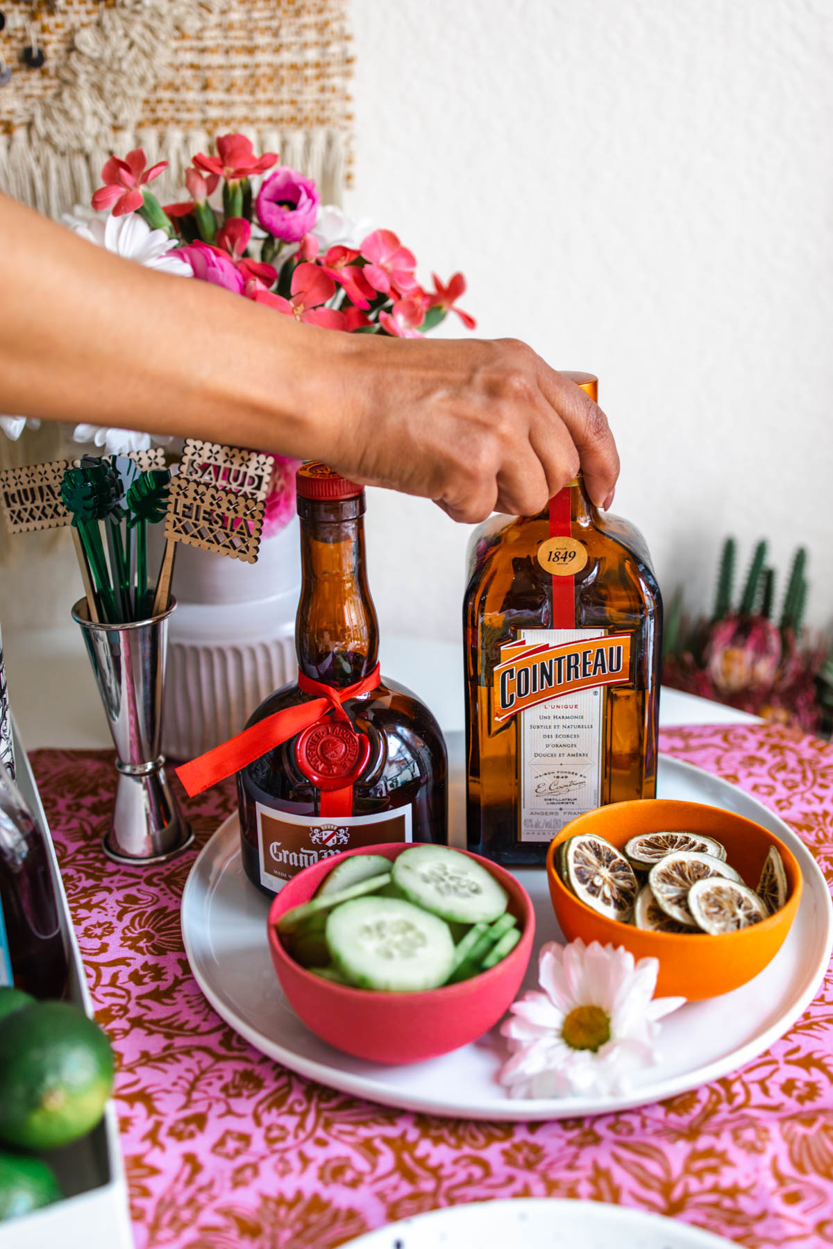 A person holding the top portion of a Cointreau bottle beside a bottle of Grand Marnier behind bowls of dried lime wheels and cucumber slices.