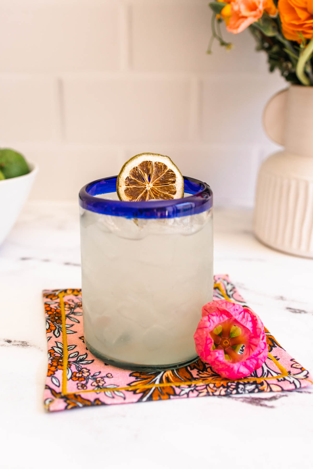 A blue-rimmed glass of Seedlip Margarita garnished with a dehydrated lime wheel on top with a pink flower below the glass.