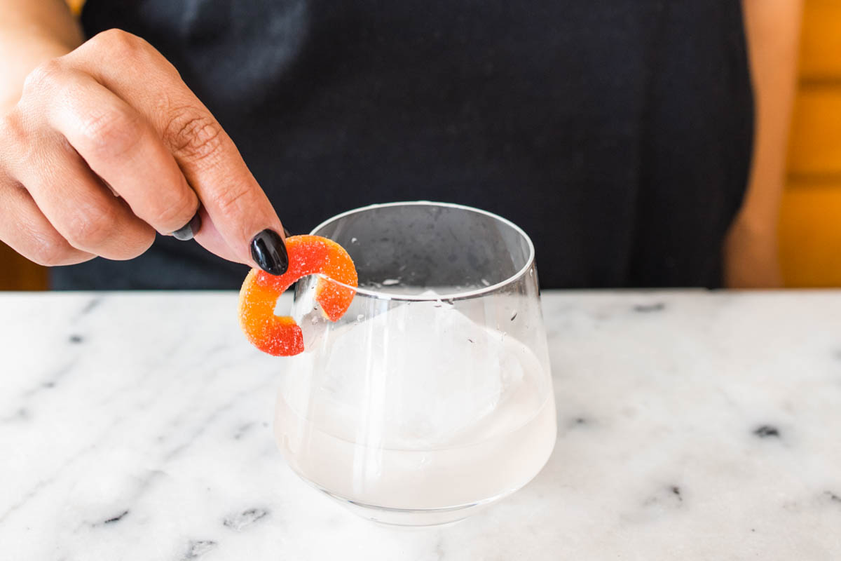 A person placing a peach candy ring at the rim of a clear glass with peach margarita.