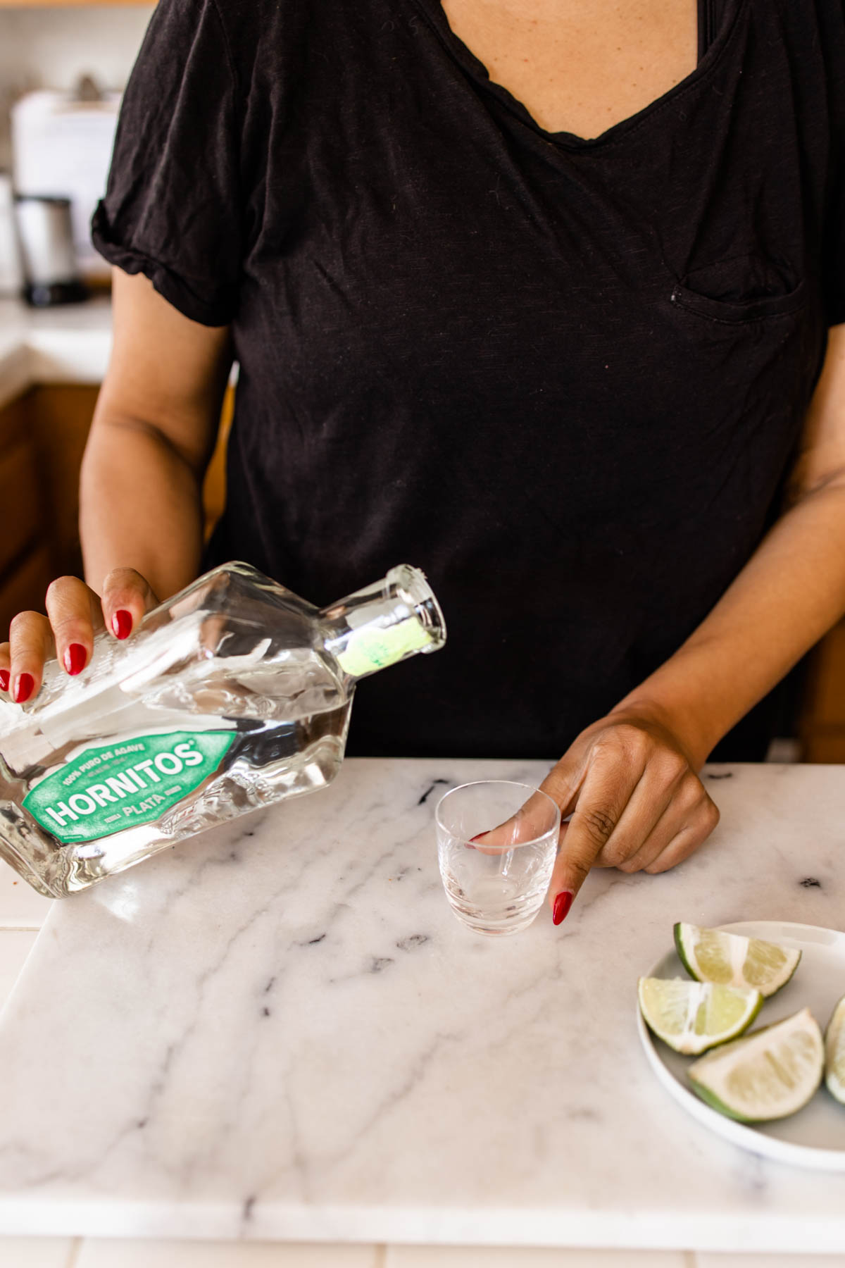 An individual that is about to pour Hornitos Tequila Plata from its bottle into an empty shot glass.