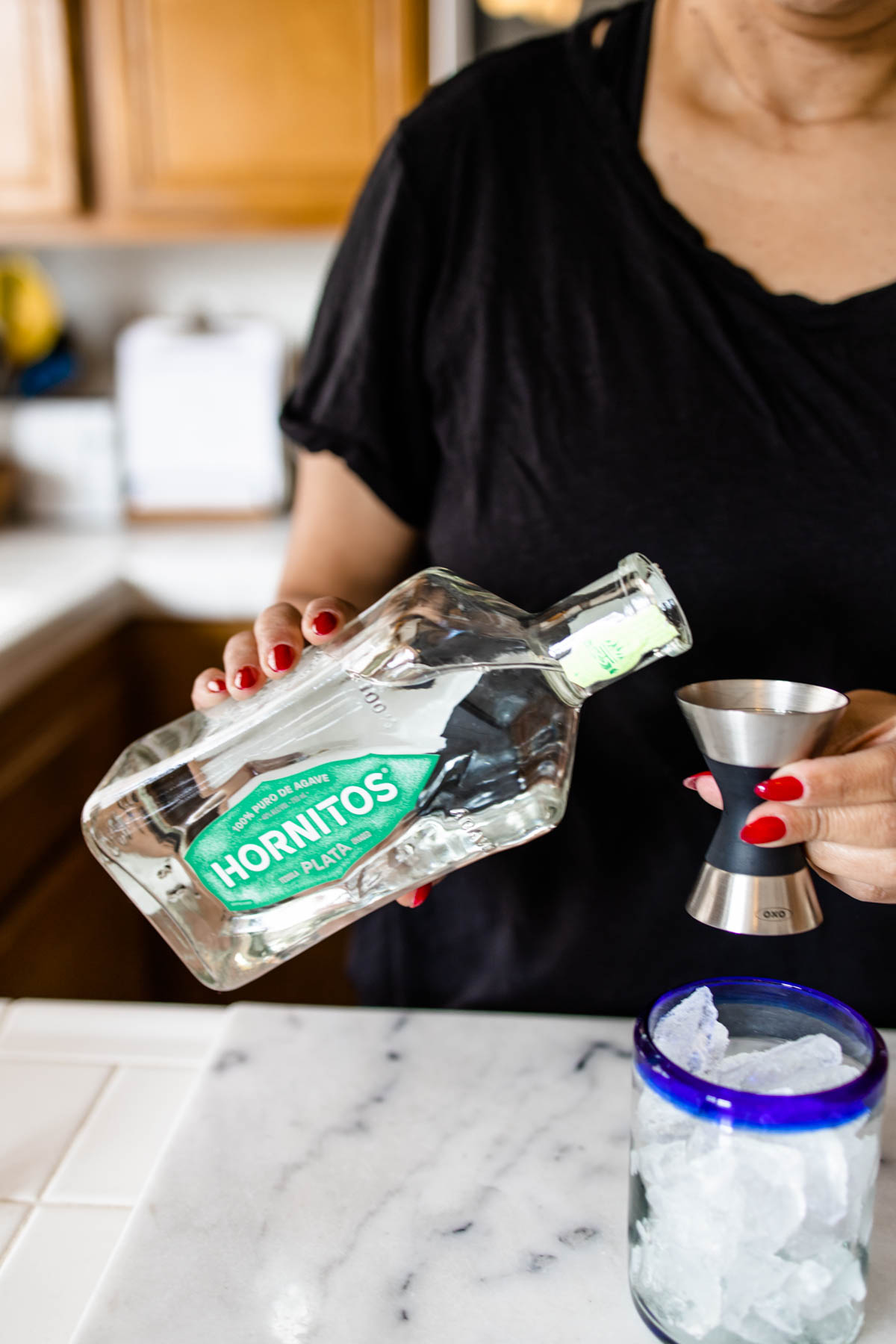 A person tilting a bottle and about to pour some Hornitos Tequila from its bottle into a jigger on top of a blue-rimmed glass with ice.