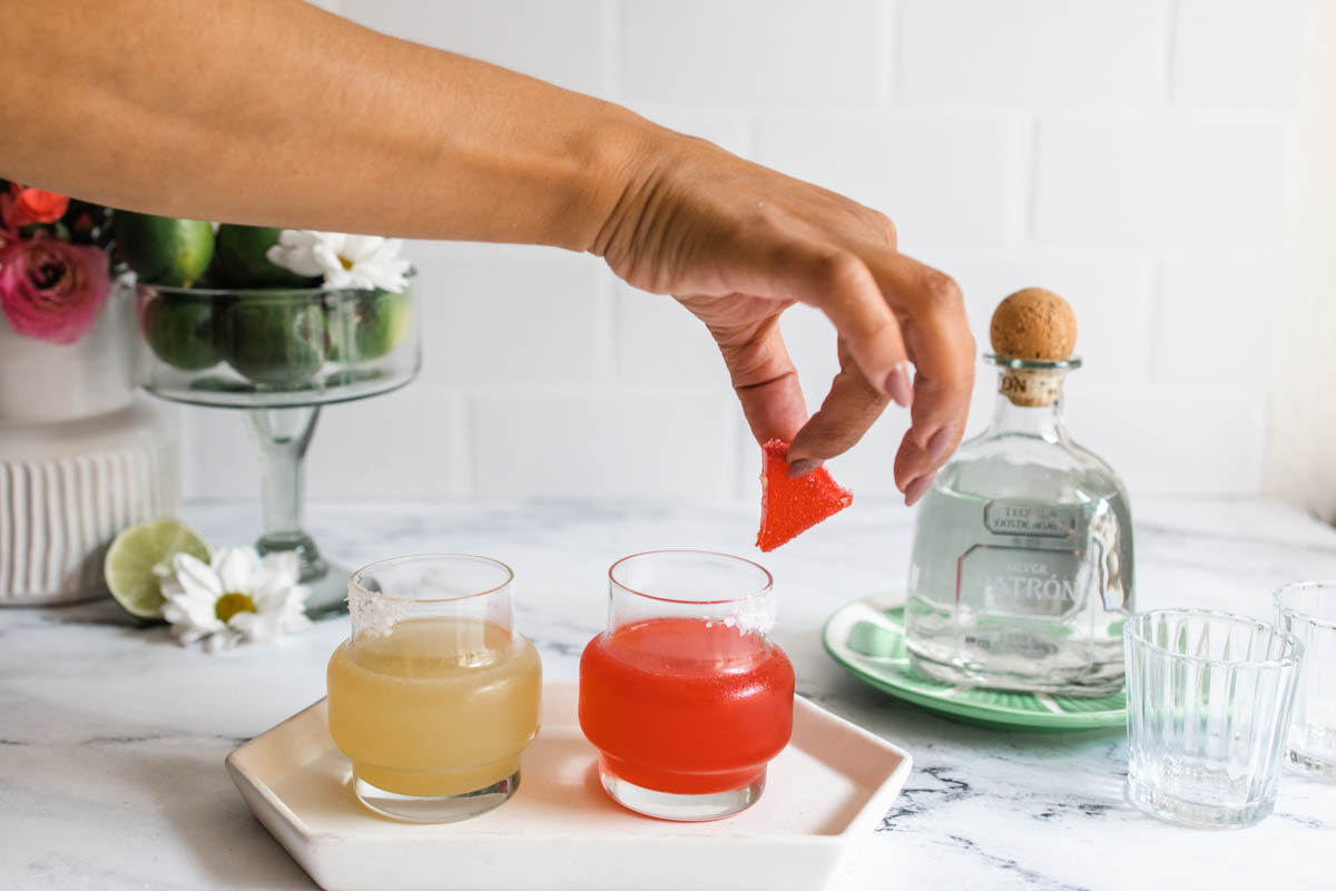 A person placing a watermelon slice on the side of a red Deadpool margarita beside a yellow Wolverine margarita on a plate.