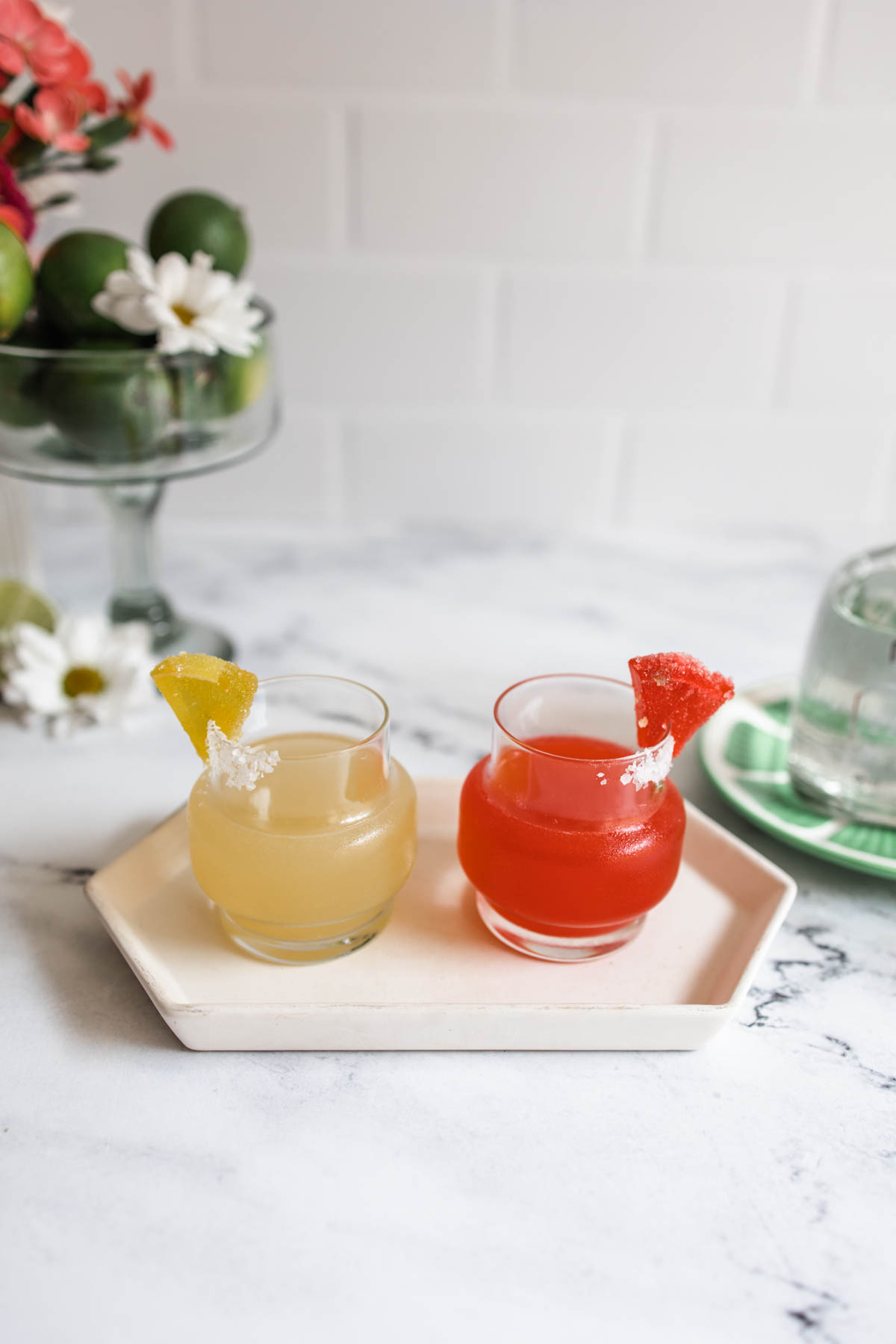 Small glasses of yellow Wolverine margarita and red Deadpool margarita on top of a white plate on a marbled countertop. 