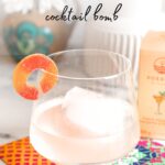 A clear glass of peach margarita with ice garnished with peach candy ring with the text "Peach Margarita Cocktail Bomb" at the top.