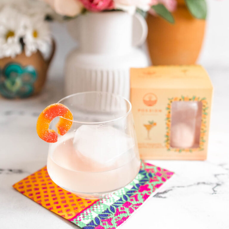 A glass of peach margarita with ice on top of a colored napkin beside a box of peach margarita drink bombs.