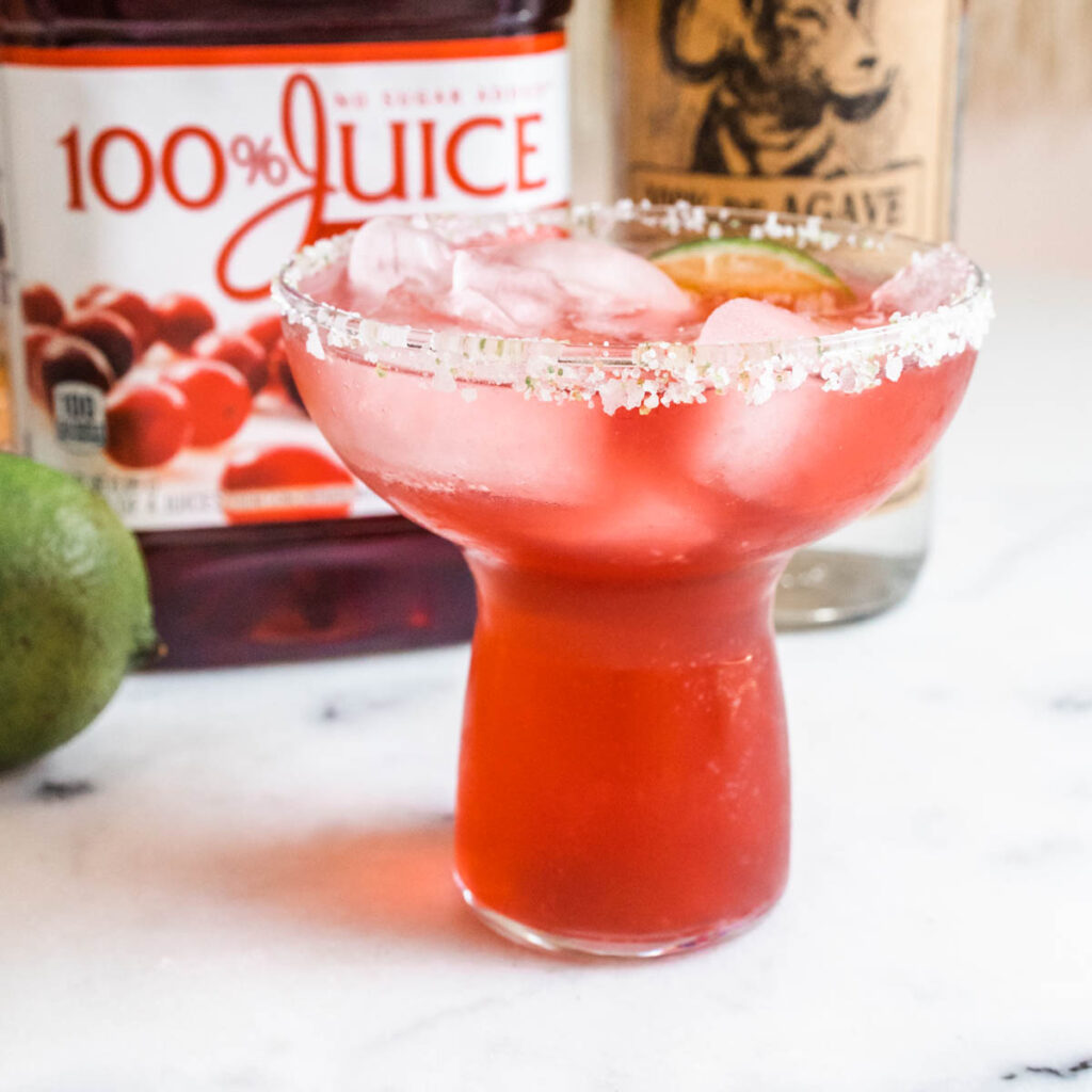 A stemless margarita glass with ice and a salted rim in front of a cranberry juice bottle.